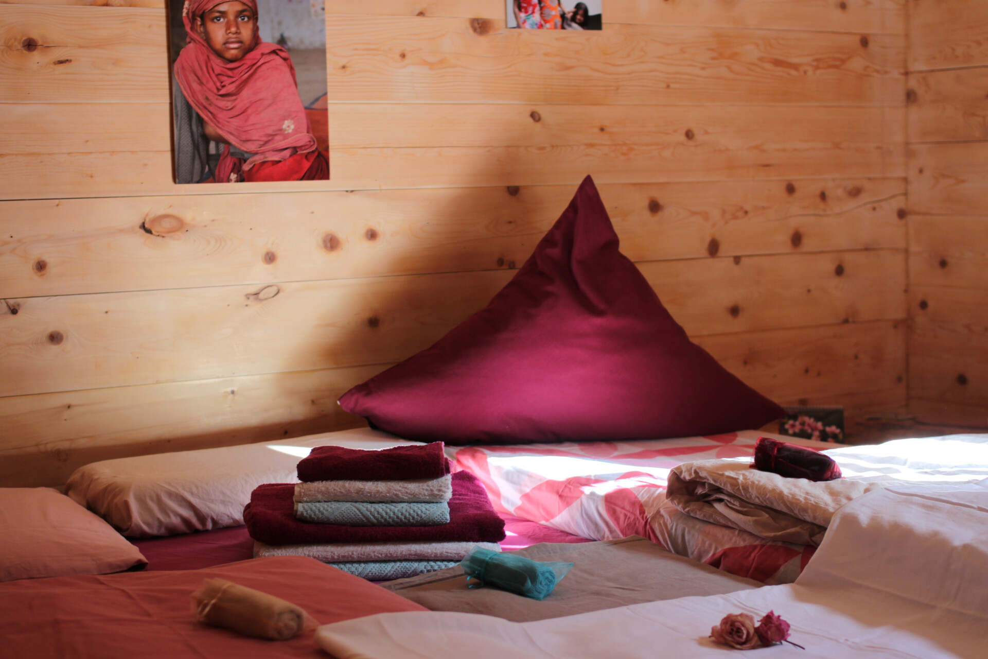 Bett mit großem rotem Polster und gemusterter Bettwäsche, bunte gefaltete Handtücher im Vordergrund